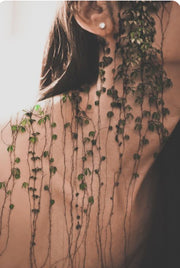Person with long hair and earrings standing behind a plant against a neutral background