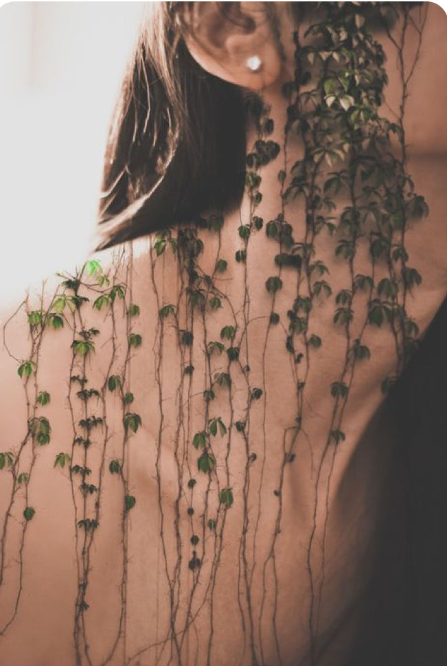Person with long hair and earrings standing behind a plant against a neutral background